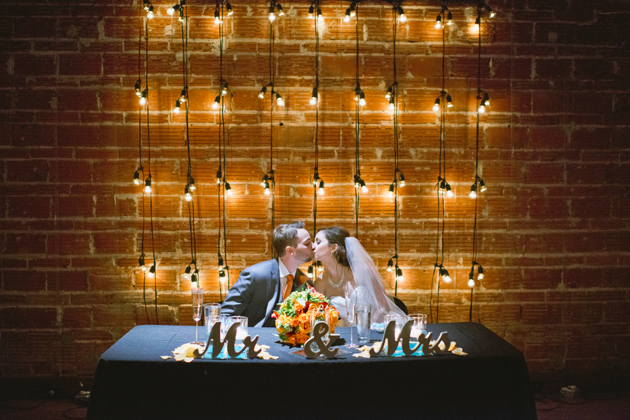 Bride and Groom Kissing at Sweetheart Table with Brick Walls and String Light Backdrop | Downtown St. Pete Wedding Venue NOVA 535 Event Space