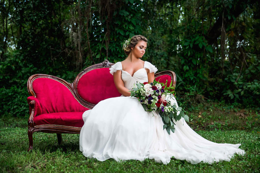 Bride in Amsale Wedding Dress from Blush Bridal Sarasota with Deep Red Burgundy Wedding Bouquet with Greenery on Red Vintage Couch Chaise | Southern Inspired Outdoor Wedding Reception Decor Styled Shoot