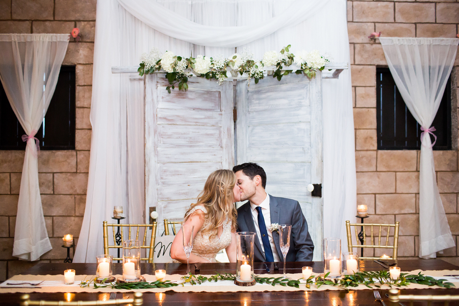 Bride and Groom Kissing at Sweetheart Table with Farm Table, Greenery and Gold Chiavari Chairs