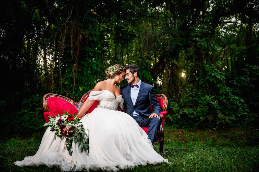 Bride in Amsale Wedding Dress from Blush Bridal Sarasota with Deep Red Burgundy Wedding Bouquet with Greenery on Red Vintage Couch Chaise | Southern Inspired Outdoor Wedding Reception Decor Styled Shoot