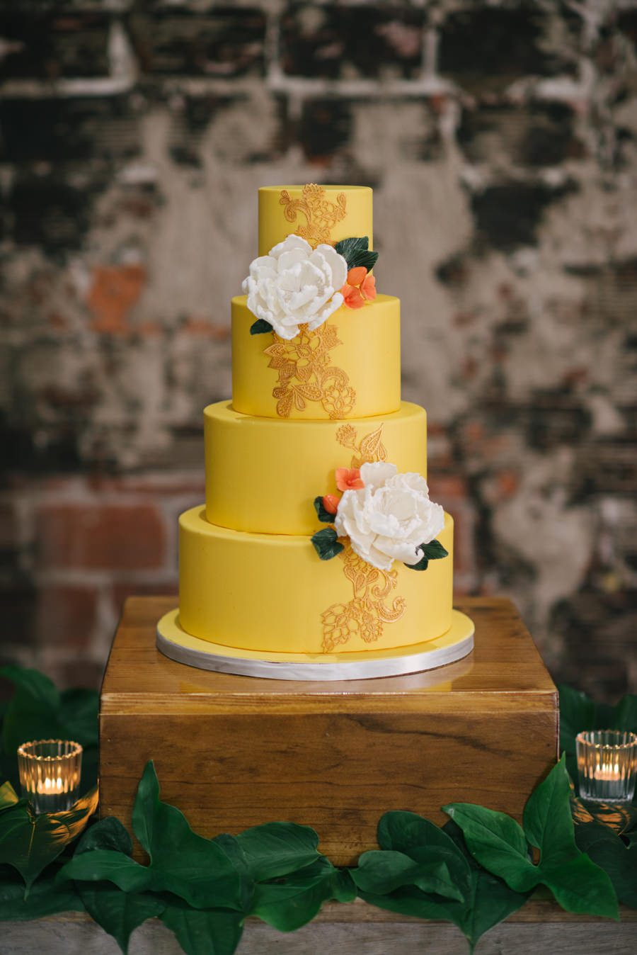 Caribbean Inspired, 4- Tiered Round Wedding Cake on Wooden Table with Yellow Fondant and Gold Design Detailing Accented with White Flowers
