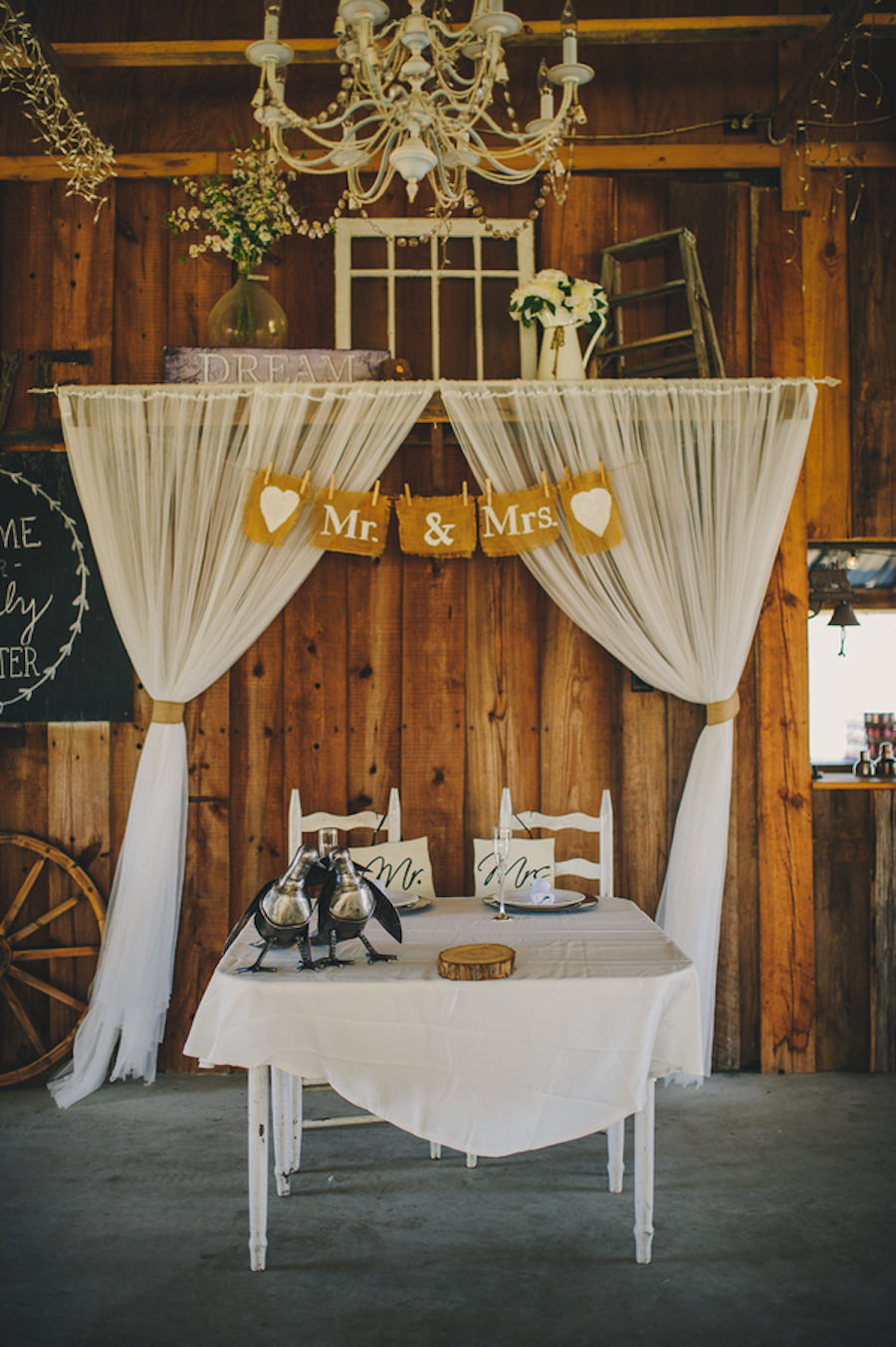 Plant City, Indoor Barn Wedding Reception Sweetheart Table with Wooden Table and Chairs, Draped Ivory Fabric Backdrop Draping and Burlap Mr and Mrs Sign