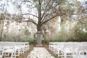Rustic Outdoor Wedding Ceremony with Light Pink and White Floral Archway under Tall Pines with White Resin Folding Chairs and Rose Petal Aisle | Tampa Bay Wedding Planner Glitz Events