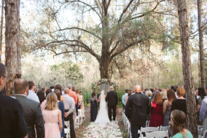 Rustic Outdoor Wedding Ceremony with Light Pink and White Floral Archway under Tall Pines with White Resin Folding Chairs and Rose Petal Aisle in Woods | Tampa Bay Wedding Planner Glitz Events