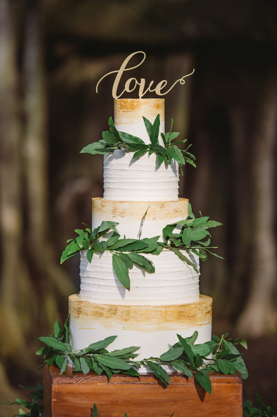 Four Tier Round White Rustic Wedding Cake with Hand Painted Gold Detail and Greenery on Wooden Cake Stand