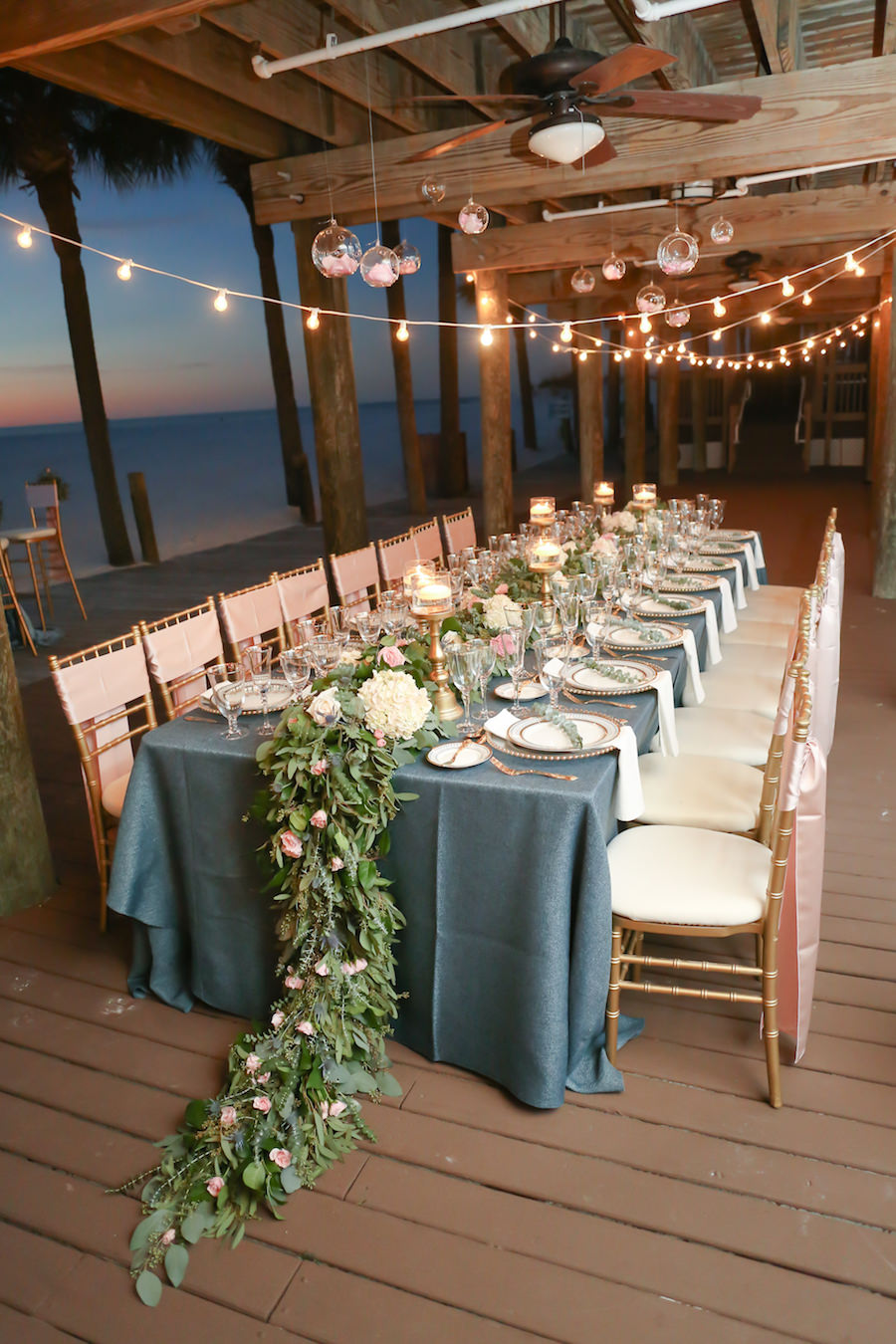 Bohemian Glam Beach Wedding with Navy Blue Burlap Linens and Floral Garland Centerpieces on Long Feasting Tables at Clearwater Beach Wedding Venue Hilton Clearwater Beach | Linens by Over The Top Rental Linens | China/Glassware by A Chair Affair | Gold Chiavari Chairs and Beaded Glass Chargers from Signature Event Rentals