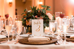 Wedding Reception Table Decor with Coral and Green Floral Centerpieces, Gold Foil Table Numbers, and Wicker Chargers