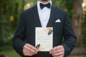 Groom in Black Tuxedo Holding Custom Ivory and Green Wedding Invitation With Magnolia Tree Floral Detail | Sarasota Wedding Stationery Citrus Press Co.