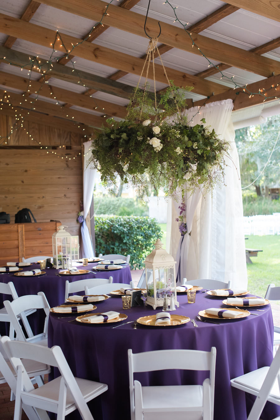 White Lantern Wedding Centerpieces with Gold Chargers on Purple Linens with White Resin Folding Chairs and Hanging Greenery Centerpiece Suspended from Ceiling | Tampa Bay Wedding Venue Cross Creek Ranch