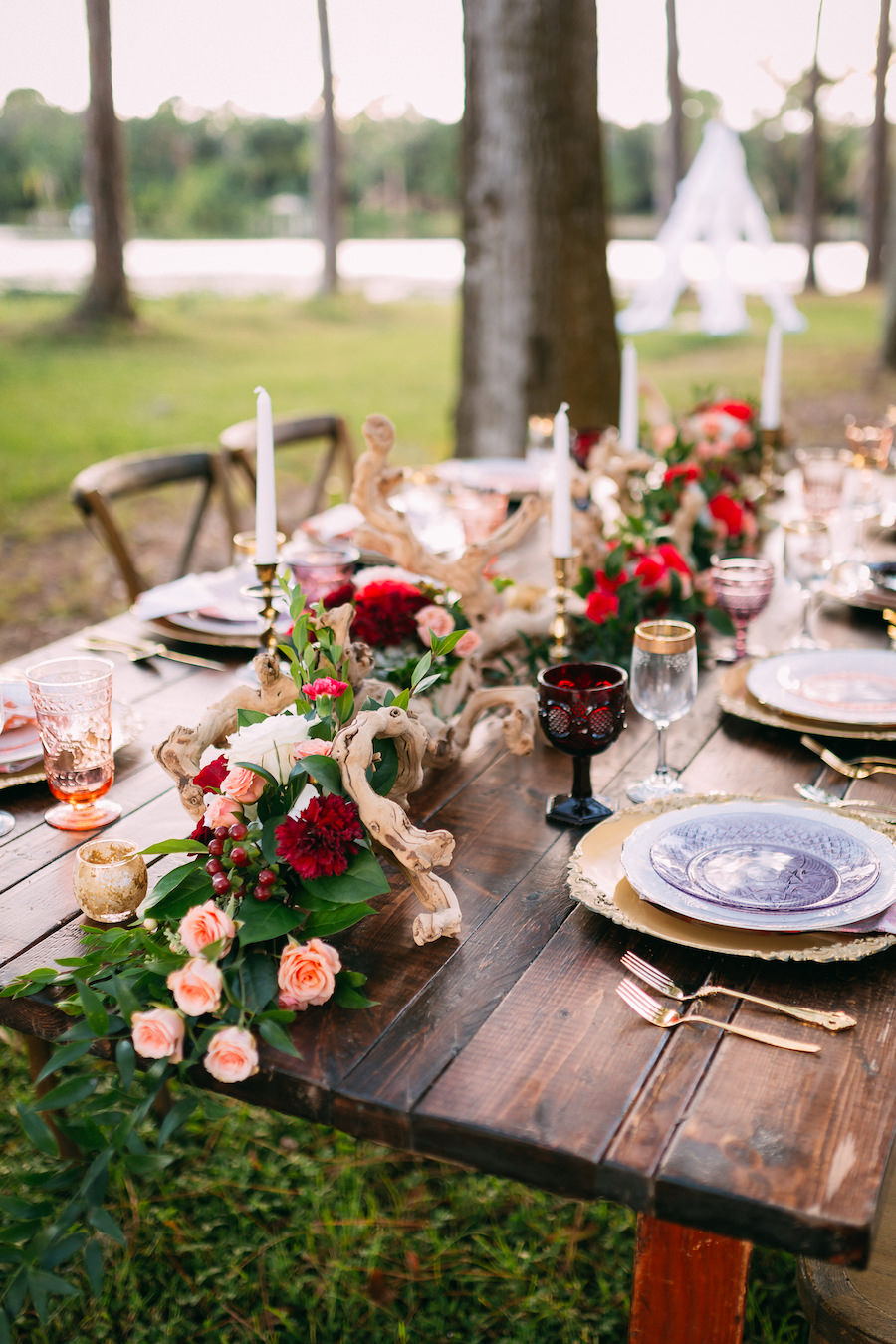 Boho Chic Wedding Reception Placesetting | Long Farm Table Feasting Table with Boho Chic Inspired Garland with Wooden Centerpiece and Vintage Glasses, Chargers and Cross Back Farm Chairs | Ever After Vintage Weddings Tampa Bay Wedding Rental Company