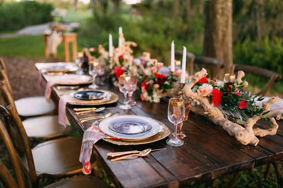 Boho Chic Wedding Reception Placesetting | Long Farm Table Feasting Table with Boho Chic Inspired Garland with Wooden Centerpiece and Vintage Glasses, Chargers and Cross Back Farm Chairs | Ever After Vintage Weddings Tampa Bay Wedding Rental Company
