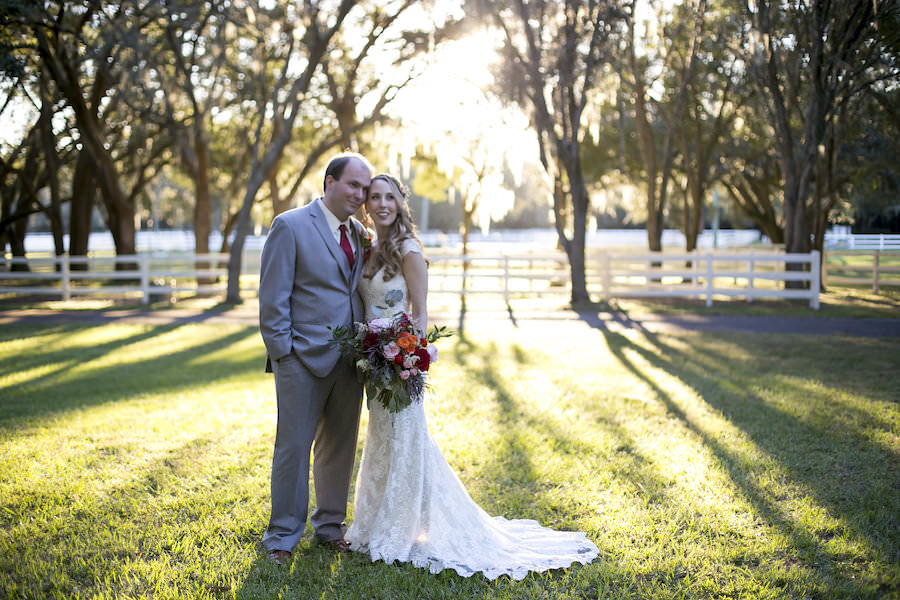 Bride and Groom Outdoor Southern Chic Wedding Portrait in Light Grey Suit and Burgundy Tie and Ivory Lace Allure Wedding Dress | Fall Themed Red Wedding Bouquet | Rustic Outdoor Wedding Tampa Bay Venue The Lange Farm