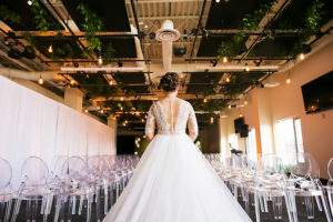 Bride Walking Down Ceremony Aisle in Nude Illusion Lace Wedding Dress with Sleeves and Clear Ghost Chairs | Tampa Bay Bridal Shop Isabel O'Neil Bridal | Rental and Decor Company A Chair Affair | Wedding Photographer Limelight Photography | Downtown Tampa Venue Glazer's Children Museum