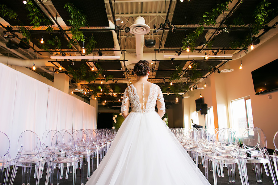 Bride Walking Down Ceremony Aisle in Nude Illusion Lace Wedding Dress with Sleeves and Clear Ghost Chairs | Tampa Bay Bridal Shop Isabel O'Neil Bridal | Rental and Decor Company A Chair Affair | Wedding Photographer Limelight Photography | Downtown Tampa Venue Glazer's Children Museum