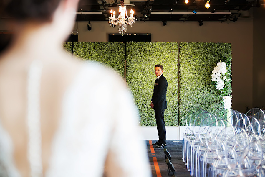 Bride Walking Down Ceremony Aisle in Nude Illusion Lace Wedding Dress with Sleeves | Greenery Hedge Wall Ceremony Backdrop with Clear Ghost Chairs and Hanging Chandelier | Tampa Bay Bridal Shop Isabel O'Neil Bridal | Rental and Decor Company A Chair Affair | Wedding Photographer Limelight Photography | Downtown Tampa Venue Glazer's Children Museum | Lighting Nature Coast Entertainment Services