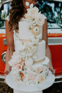 Bride Holding 3-Tier Round Hand Painted Wedding Cake with Cascading Sugar Flowers and Gold Leaf Boarder | Retro Vintage Boho Wedding Inspiration with Orange VW Bus | Tampa Wedding Cake Bakery Hands on Sweets