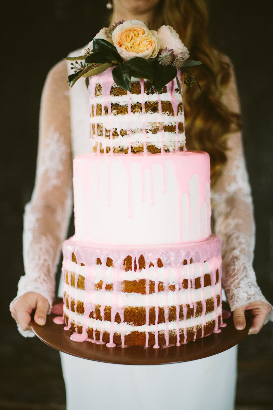 Bride in Long Sleeve Lace Isabel O'Neil Bridal Collection Wedding Dress Holding Three Tiered Round Naked Wedding Cake with Dripping Pink Icing and Fresh Rose Floral Cake Topper | Tampa Bay Wedding Planner Isabel O'Neil Bridal Collection