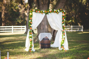 Outdoor Dade City Rustic Fall Inspired Wedding Ceremony Floral Arch Decor with Orange, Yellow Flowers, Greenery Accents, Wine Barrels and Soft Fabric Draping | The Lange Farm Wedding