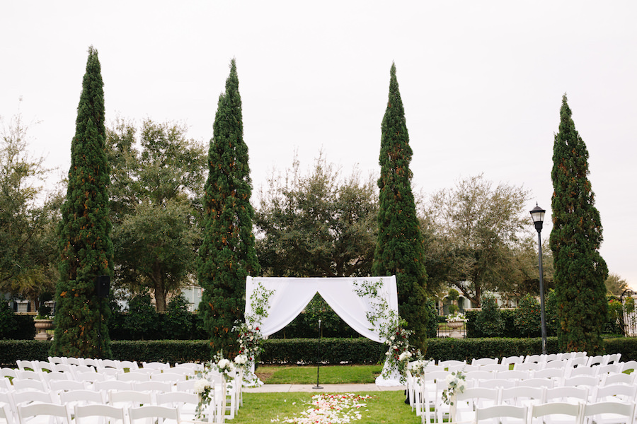 Outdoor Ceremony Decor at Tampa Bay Garden Wedding Venue The Palmetto Club with White Fabric Arch, Climbing Greenery and Red Rose Bouquets, and White Folding Chairs with Rose Petal Aisle