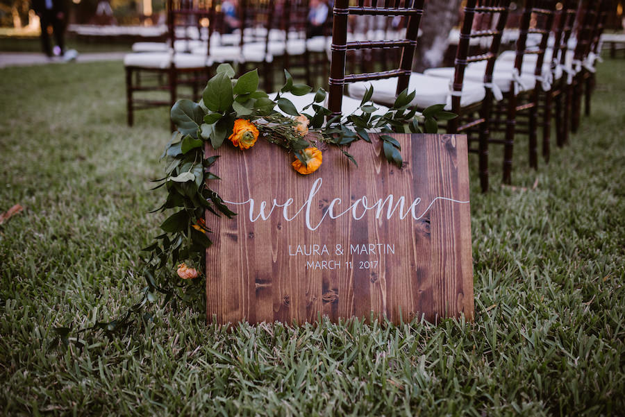 Rustic Outdoor Wedding Ceremony Painted Elegant Wooden Welcome Sign with Greenery and Orange Floral Garland and Black Chiavari Chairs with White Cushions
