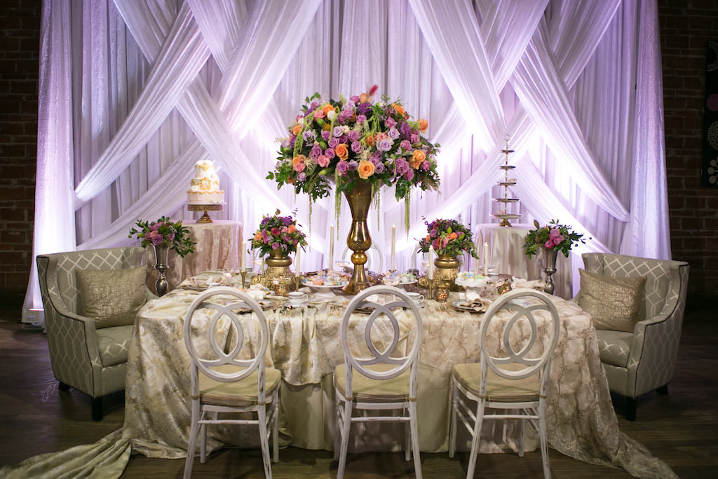 Modern Wedding Reception with Unique Vintage Inspired Furniture and Linens, Tropical Orange, Pink, and Purple Rose with Greenery Centerpieces in Tall Gold Vases, and Drapery with Purple Uplighting | St. Petersburg Wedding Venue NOVA 535 | Planner UNIQUE Weddings and Events | Photographer Carrie Wildes Photography