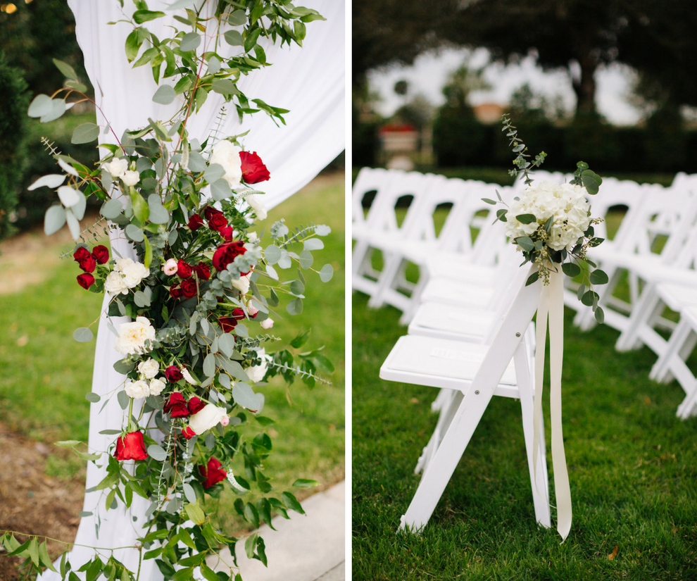 Ceremony Decor at Tampa Bay Garden Wedding Venue The Palmetto Club with White Fabric Arch with Climbing Greenery and Red and White Rose Bouquets, and White Folding Chairs with White Floral Bouquet and Cream Ribbon
