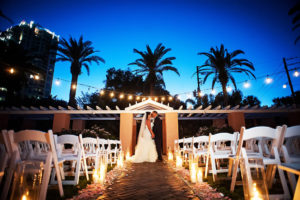 Outdoor Nighttime Courtyard Wedding Ceremony Portrait with Pink Rose Petal Aisle, String LIghts, White Folding Chairs, and Tall Hurricane Candle Holders | St Pete Wedding Photographer Limelight Photography | Tampa Bay Hotel Wedding Venue Vinoy Renaissance