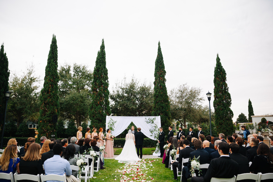 Outdoor Garden Wedding Ceremony at Tampa Bay Venue the Palmetto Club with White Fabric Arch with Climbing Greenery and Rose Petal Aisle and White Folding Chairs, Blush Bridesmaids Dresses