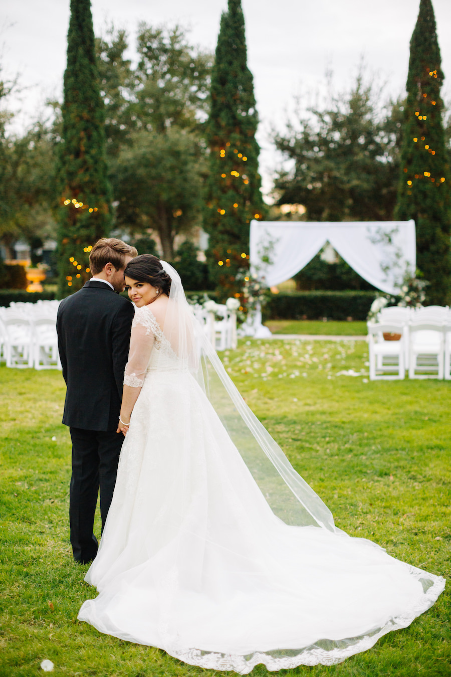 Bride and Groom Wedding Portrait at Garden Ceremony Venue The Palmetto Club with String Light Wrapped Trees, White Fabric Arch, and A Line Sophia Tolli Wedding Dress with Long Train and Veil | Tampa Bay Wedding