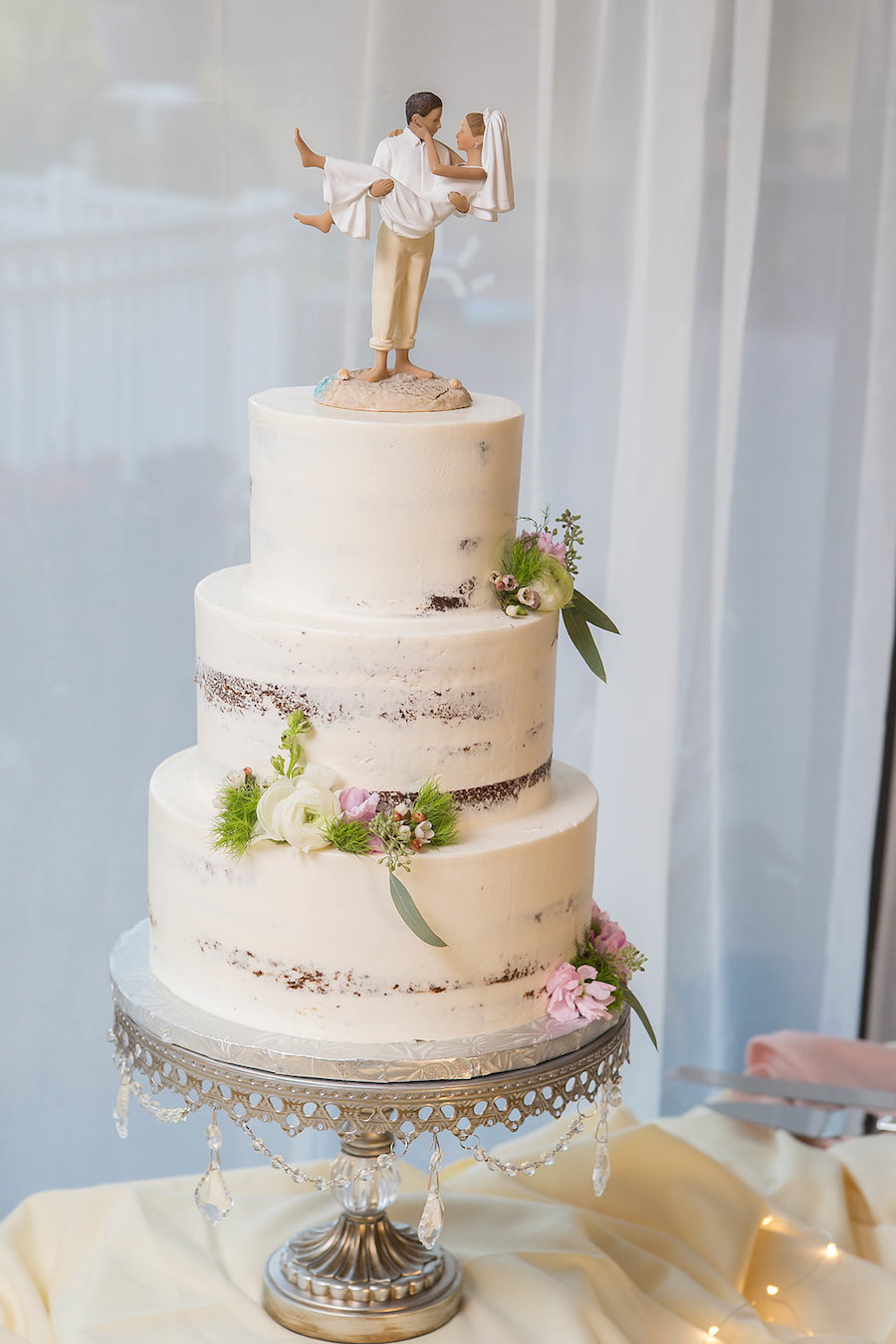Semi-Naked Three Tier Round Natural Rustic Wedding Cake with Beach Inspired Florals and Playful Beach Wedding Bride and Groom Figurine Cake Topper on Silver Jeweled Cake Stand | Tampa Bay Wedding Bakery The Artistic Whisk