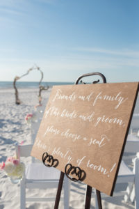 Hand-painted Wooden Welcome Sign at Beach Wedding Ceremony with Natural Driftwood Arch, Folding White Chairs with White and Pink Flowers | St Petersburg Wedding Venue The Don Cesar