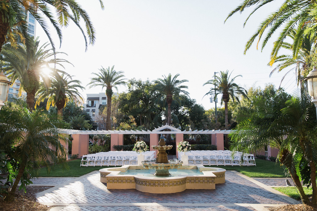 Outdoor Hotel Garden Courtyard Wedding Ceremony 1920s Art Deco Venue THe Vinoy Renaissance with Fountain, Large Floral Arrangements on Pedestals, and White Folding Chairs | Tampa Bay Wedding Venue The Vinoy Renaissance