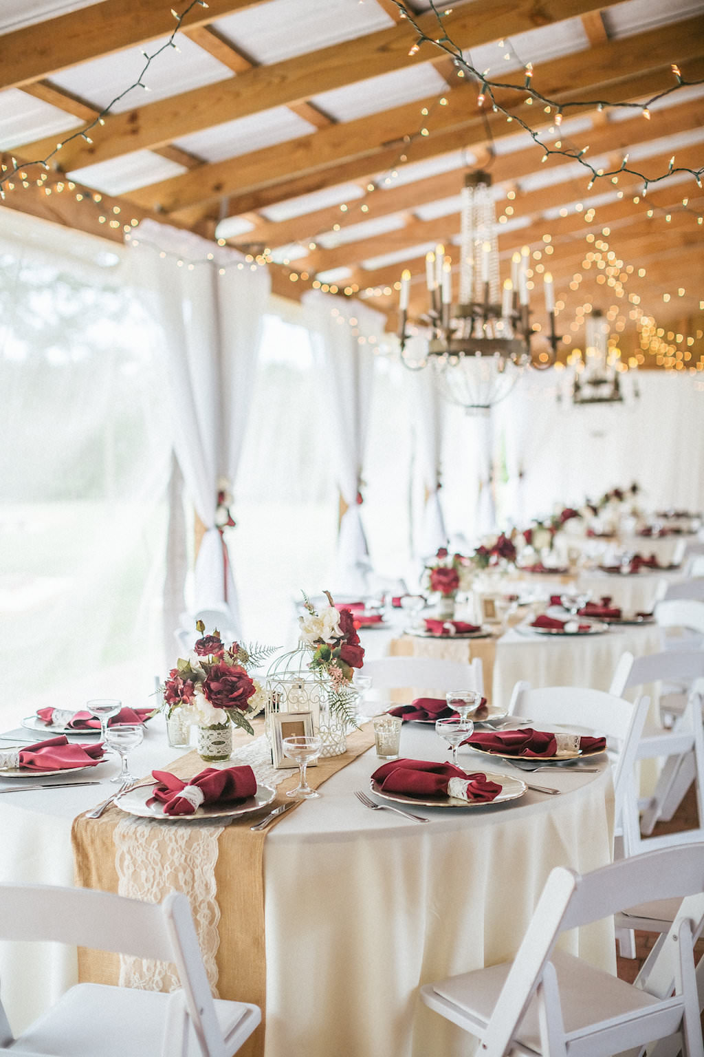 Rustic Barn Wedding Reception with String LIghts, Chandeliers, Burlap and Lace Runners on Cream Linen with Burgundy Napkins and Red Rose Centerpieces in Mason Jars with White Folding Chairs