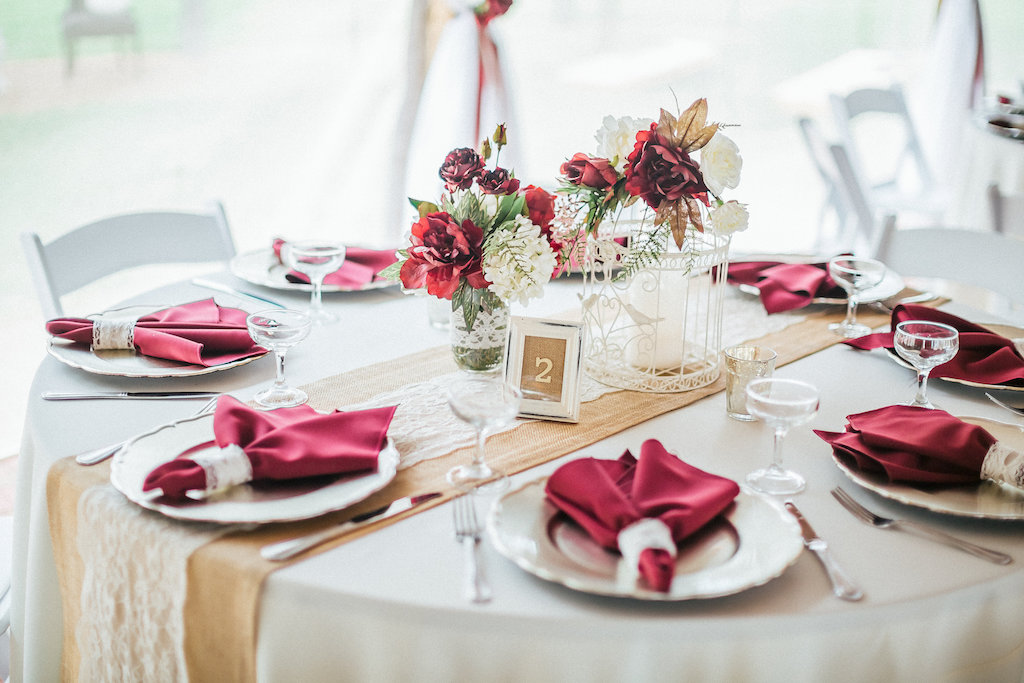 Rustic Barn Wedding Reception with Burlap and Lace Runners on Cream Linen with Marsala Napkins and Red Rose, Fall Foliage, and White Floral Centerpieces in Mason Jars and vintage Frame Table Number with White Folding Chairs