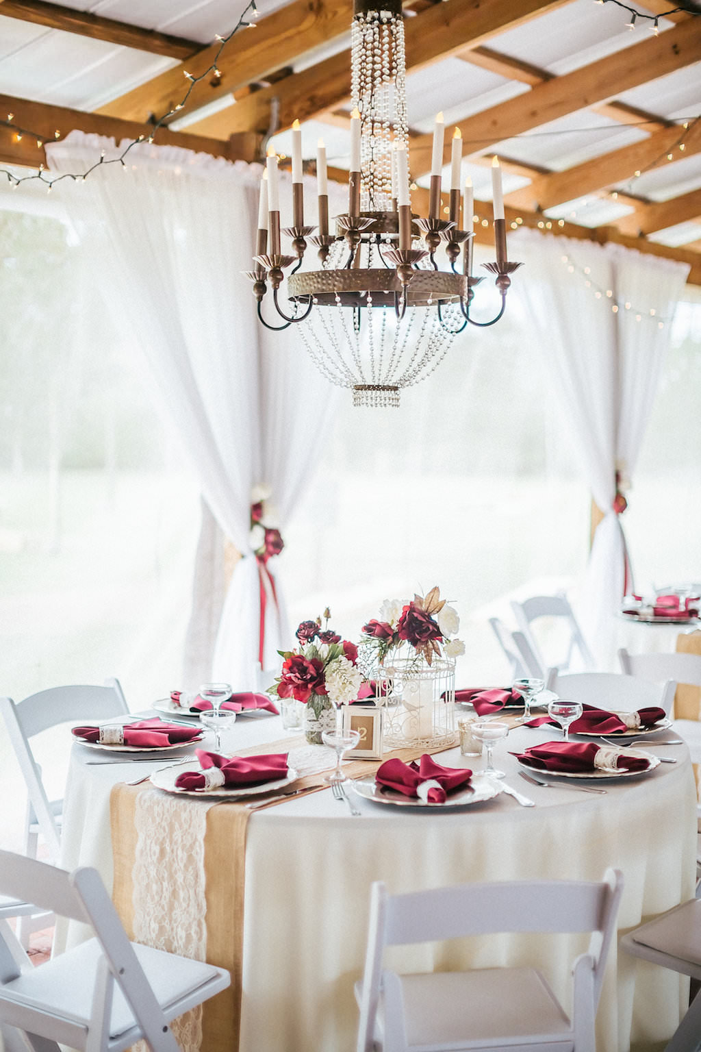 Rustic Barn Wedding Reception with String LIghts, Chandeliers, Burlap and Lace Runners on Cream Linen with Burgundy Napkins and Red Rose Centerpieces in Mason Jars with White Folding Chairs
