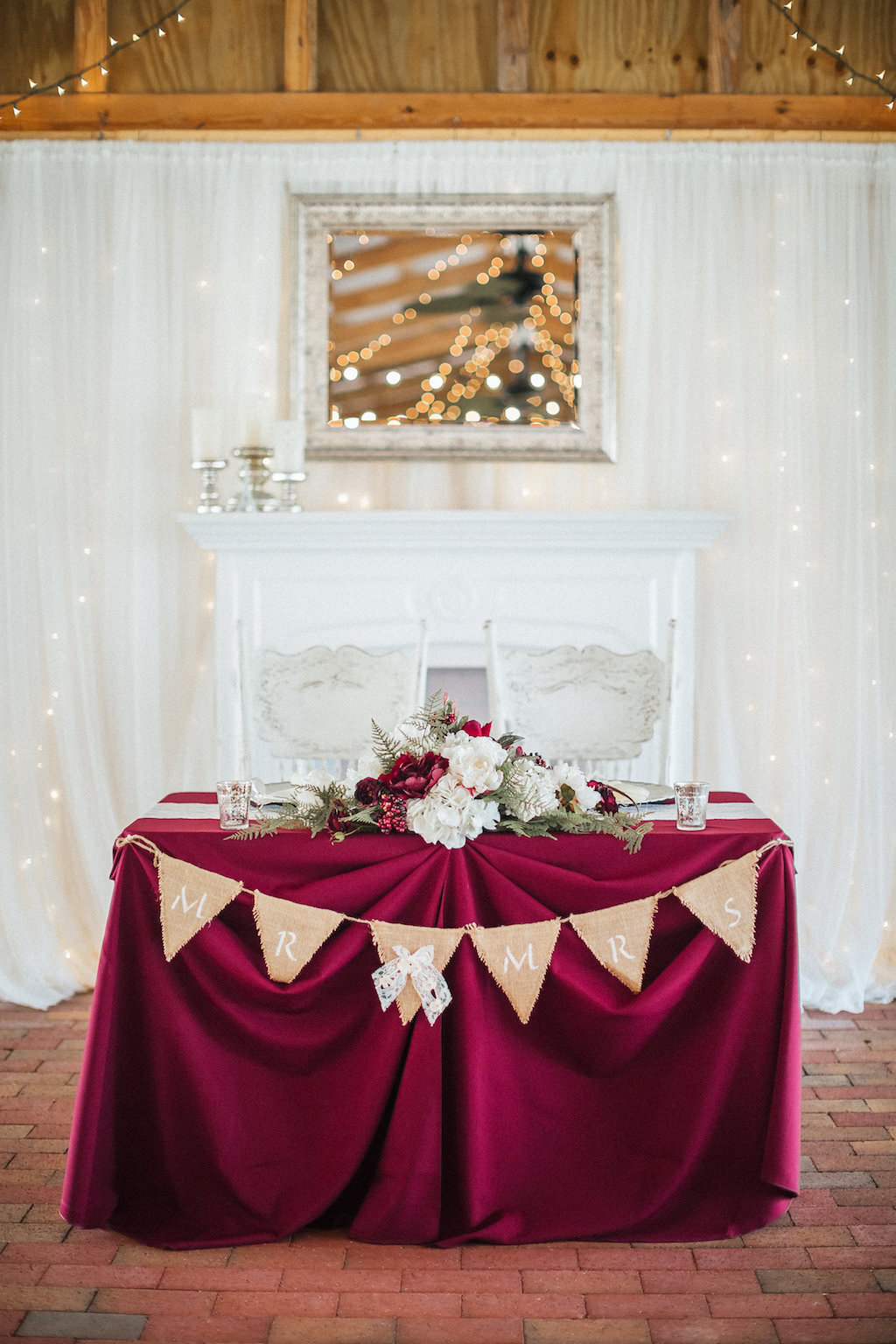 Rustic Barn Wedding Bride and Groom Sweetheart Table with Burlap Pennant Sign, Burgundy Tablecloth, White Lace Draping and String Lights, and Low Cream and Marsala with Greenery Centerpiece