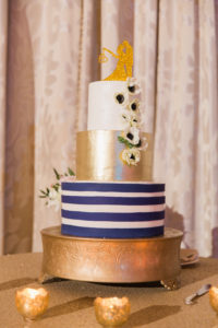 Three Tiered Round Wedding Cake with Bold White and navy Stripes, Gold Icing, and White Anemones with Bride and Groom Dancing Gold Glitter Cake Topper on Gold Cake Stand with Gold Candles