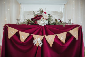 Rustic Farm Wedding Sweetheart Table with Burlap Pennant Mr and Mrs Sign on Red Tablecloth with Red Berry and White Floral with Ferns low Centerpiece