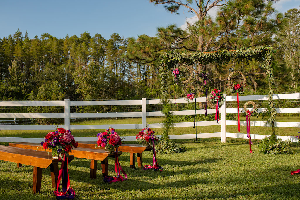Outdoor Rustic Wedding Ceremony Decor with Wooden Benches, and Small Red, Purple, and Magenta Flowers with Pink and Purple Ribbon, and Rustic Ceremony Arch with Greenery Garland and Hanging Woven Branch Wreaths with Ribbon | Tampa Bay, Florida Wedding Venue Southern Plantation Oasis | Wedding Planner Exquisite Events | Northside Florist