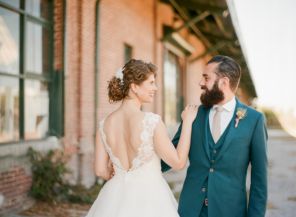 Bride and Groom Outdoor Industrial Wedding Portrait, Bride in V Back Lace Paloma Blanca Wedding Dress, Groom in Teal Suit with Tropical Yellow and Pink Boutonniere and Gray Tie
