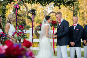 Outdoor Southern Wedding Ceremony Portrait with Greenery Garland Arch, and Hanging Woven Branch Wreaths with Purple, Magenta, and Red Roses with Ribbon | Groomsmen in Navy Suit Jackets in Hot Pink Bow Ties with White Pants | Tampa Bay Wedding Photographer Caroline & Evan Photography | Lutz Florida Wedding Venue Southern Plantation Oasis | Northside Florist | Wedding Planner Exquisite Events