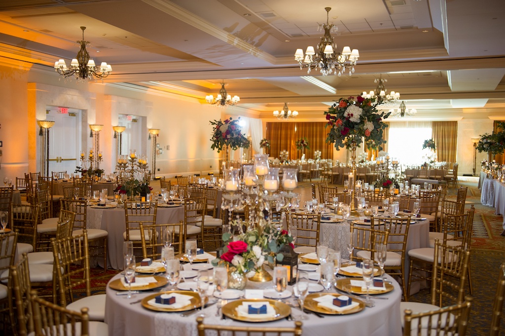 Old Florida Inspired Ballroom Wedding Reception with Navy Blue Favors, Gold Chargers and Chiavari Chairs, and Tall Gold and Glass Candle Lantern Antique Candelabra Red Rose with White Hydrangea and Greenery Centerpiece | Downtown Tampa Hotel Wedding Venue Tampa Marriott Waterside