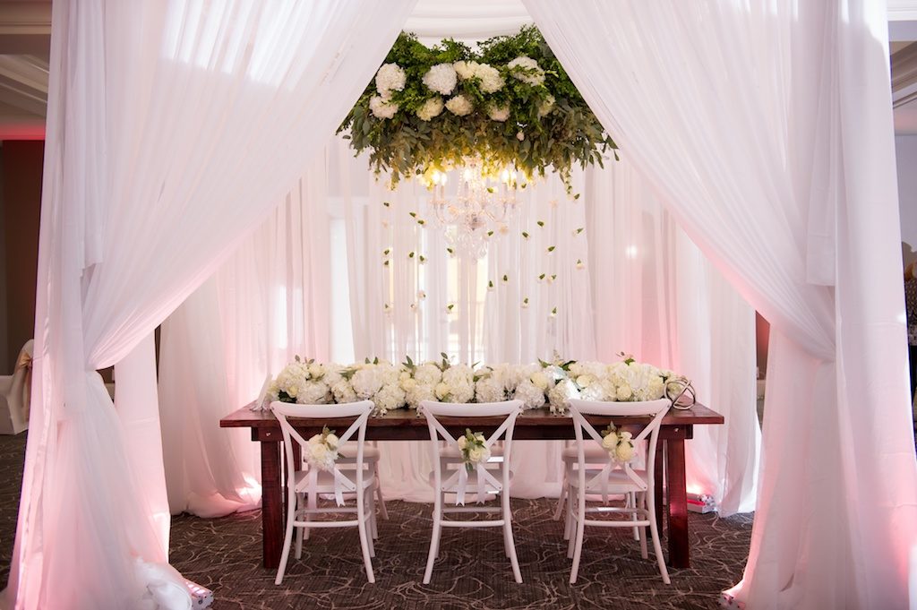 Elegant Southern White Wedding Reception Decor with Suspended White Florals with Hanging Organic Greenery, Low White Hydrangea Table Runner Garland Centerpiece, and White X Back Chairs with White Flowers and Ribbon at Wooden Farmhouse Table | Tampa Bay Wedding Planner Love Lee Lane | Tampa Event Rentals and Florist Gabro Event Services | Wedding Rentals A Chair Affair