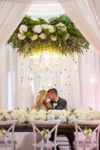 Elegant Southern White Wedding Reception Bride and Groom Portrait with Suspended White Florals with Hanging Organic Greenery, Low White Hydrangea Table Runner Garland Centerpiece, and White X Back Chairs with White Flowers and Ribbon at Wooden Farmhouse Table | Tampa Bay Wedding Planner Love Lee Lane | Tampa Event Rentals and Florist Gabro Event Services | Wedding Rentals A Chair Affair | Tampa Wedding Photographer Andi Diamond