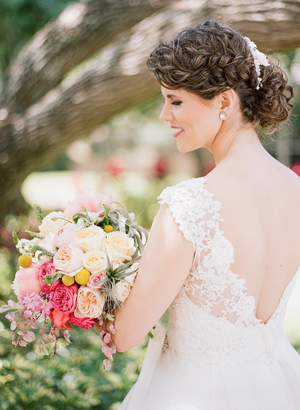 Outdoor Bridal Portrait wearing Lace Sleeve Open V Backed Paloma Blanca Wedding Dress with Peach, Blush, Magenta, and Yellow Bouquet with Greenery with Stylish Side Braid Wedding Updo Hair