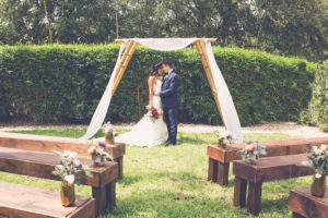 Natural Outdoor Garden Wedding Ceremony Bride and Groom Portrait with Magenta and Peach Rose with Greenery Bouquet, Sage Green Flower Arrangements in Gold Geodesic Vases on Rustic Wooden Pews, with Bamboo Ceremony Arch with White Draping | Tampa Bay Wedding Ceremony Venue Crowne Plaza Tampa