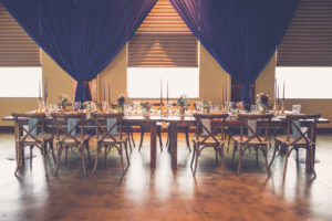 Natural Rustic Inspired Navy and Bordeaux Hotel Wedding Reception with Blue Draping, Wooden X Back Chairs, Long Wooden Tables, Low Floral Centerpieces and Tall Candles | Tampa Bay Modern Wedding Reception Venue Crowne Plaza Tampa | Tampa Farm Table Rentals A Chair Affair