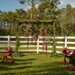 Outdoor Rustic Wedding Ceremony Decor with Wooden Benches, and Small Red, Purple, and Magenta Flowers with Pink and Purple Ribbon, and Rustic Ceremony Arch with Greenery Garland and Hanging Woven Branch Wreaths with Ribbon | Tampa Bay, Florida Wedding Venue Southern Plantation Oasis | Wedding Planner Exquisite Events | Northside Florist