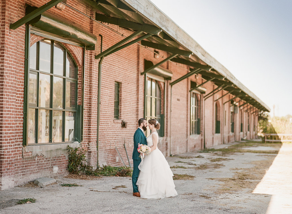 Bride and Groom Outdoor Industrial Wedding Portrait in Downtown St. Pete, Bride in Paloma Blanca Cathedral Train Wedding Dress with Coral Flower Bouquet, Groom in Teal Suit with Tropical Yellow and Pink Boutonniere