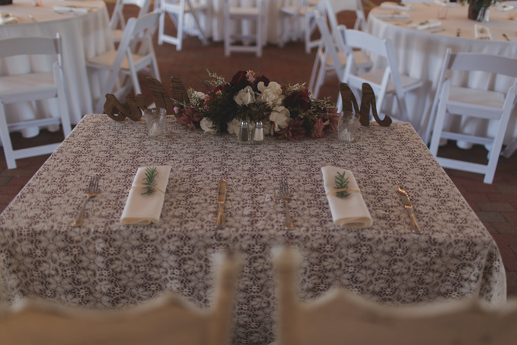 Rustic Barn Wedding Reception with Round White Tables and Folding Chairs, Sweetheart Table with White, Dusty Rose, and Maroon Centerpiece and Stylish Mr and Mrs Letters, and Rosemary and Twine Napkins on Vintage Floral Lace Linen